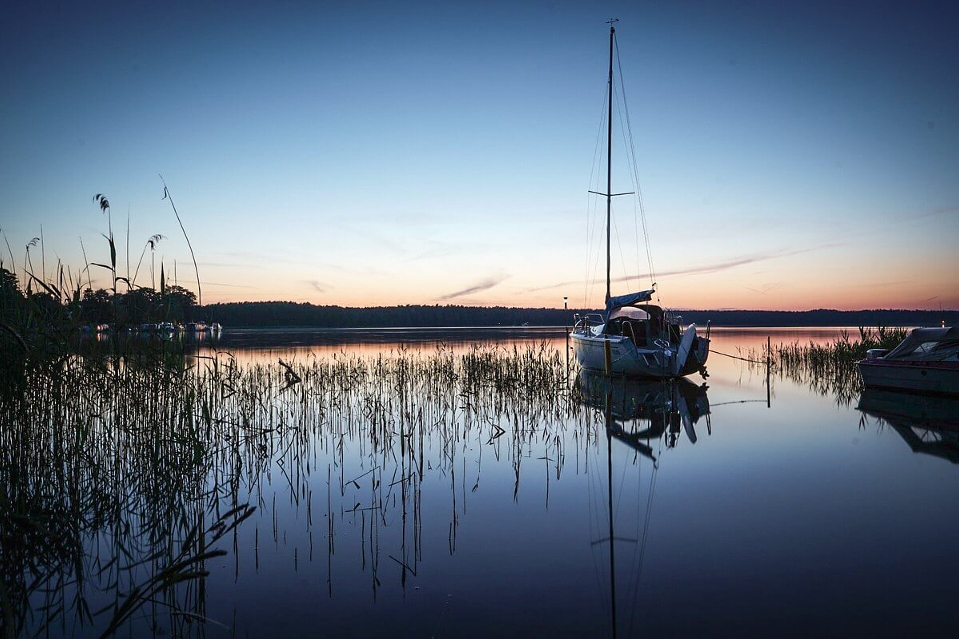 Ein Segelboot liegt bei Sonnenuntergang am Rand eines spiegelglatten Sees. Drumherum steht Schilf.